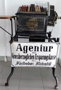 eine Addiermaschine für Banken aus dem Jahr 1900 - davor ein Email-Schild: Agentur der Grossherzoglichen Ersparungskasse des Fürstentums Birkenfeld